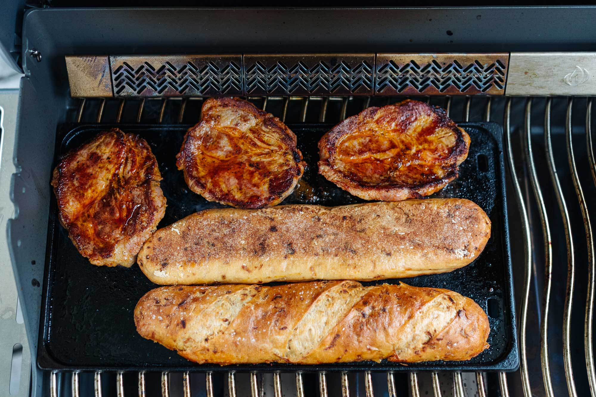 Auf einem Grill wird das rustikale Baguette kurz angeröstet. Es liegen Schweinenackensteaks mit im Grill