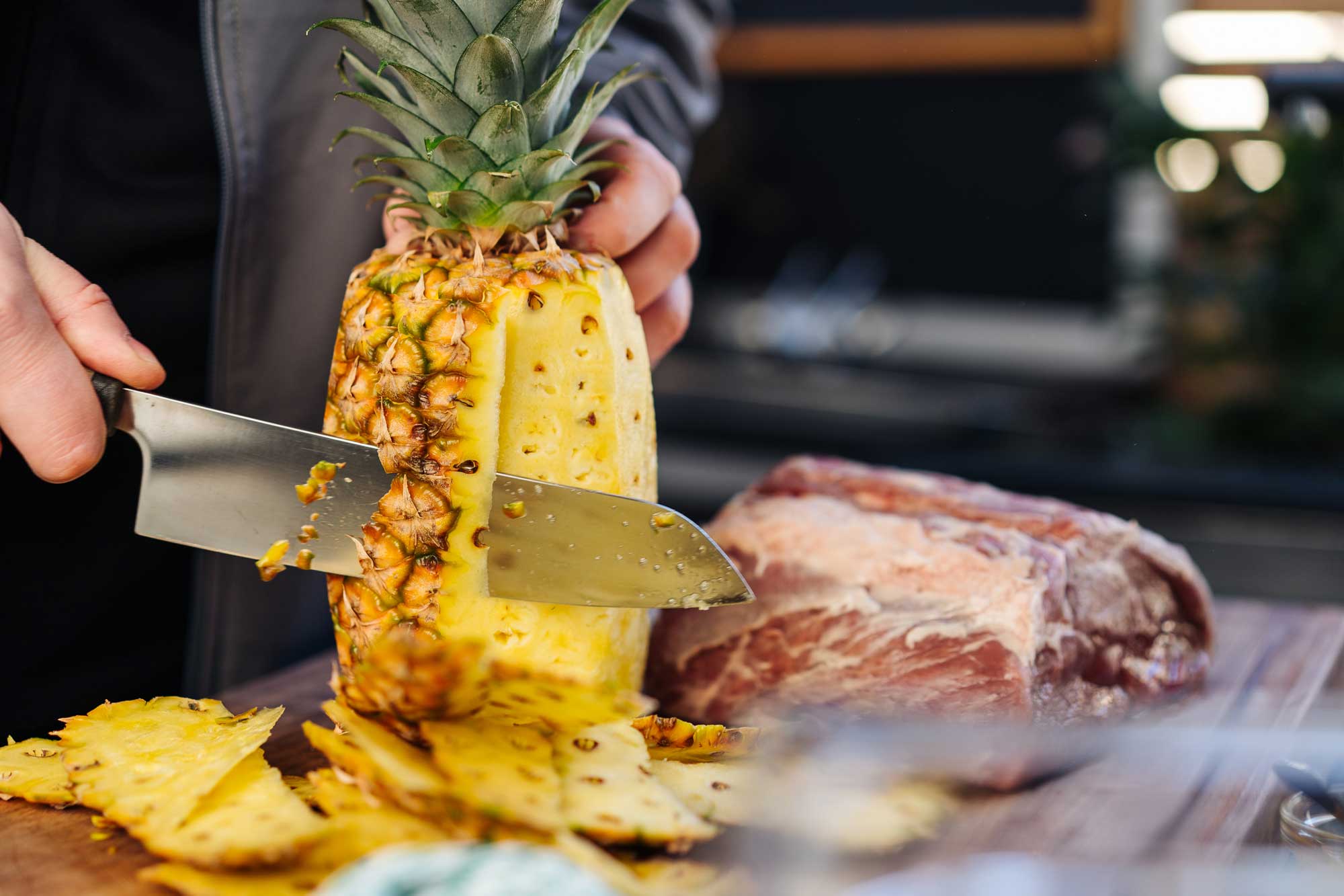 Von der Ananas wird die Schale heruntergeschnitten. Daneben liegt ein roher Rib Eye Braten